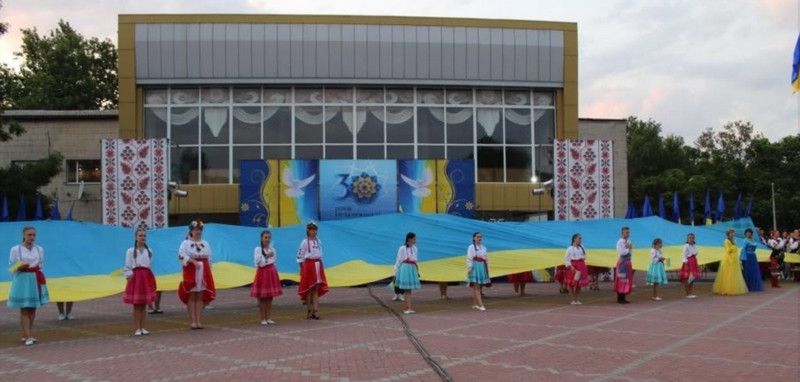 Дівчата й хлопці в українському традиційному вбранні розгортають великий синьо-жовтий прапор на площі перед будинком культури на тлі банера «30 років Незалежності».