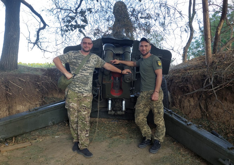 Двоє військовослужбовців у камуфляжній формі стоять біля артилерійської гармати, розміщеної в окопі серед дерев.