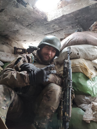 Український військовий у захисному шоломі, камуфляжній формі та рукавицях сидить у бліндажі або укріпленому окопі, укріпленому мішками з піском. Він посміхається, а поруч із ним стоїть автоматична зброя. 