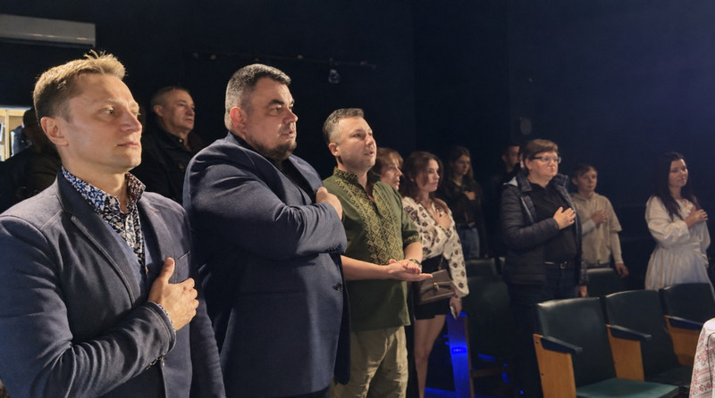 Група людей стоїть у залі між рядами крісел. Більшість тримає праву руку на грудях і дивиться вперед. Серед присутніх — чоловіки в піджаках і вишитій сорочці та жінки у вишиванках і повсякденному одязі. 