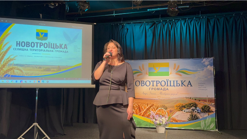 Жінка в темній сукні виступає з мікрофоном на сцені на тлі банера та екрана із символікою Новотроїцької громади. Поруч на сцені стоїть кошик із квітами.