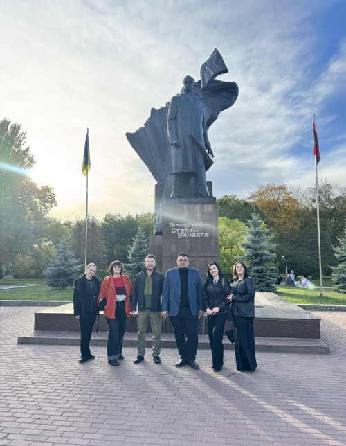 Група людей позує перед пам’ятником із написом «Герой України Степан Бандера» на площі. Позаду — дерева, клумби та прапори. 