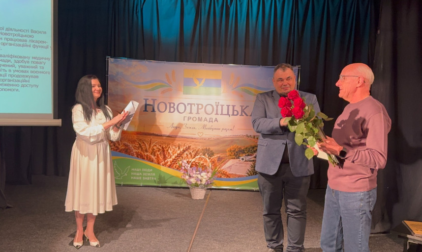 Нагородження на сцені: праворуч стоять два чоловіки, один з яких тримає букет троянд, які йому вручили з нагоди вручення подяки, а ліворуч стоїть ведуча у світлій сукні з паперами в руках. На задньому плані розташований баннер із написом «Новотроїцька громада»