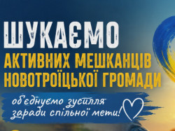 Плакат із закликом до єдності мешканців Новотроїцької громади на тлі заходу сонця та складених разом рук. Угорі розміщено синьо-жовте серце та заголовок: «ШУКАЄМО АКТИВНИХ МЕШКАНЦІВ НОВОТРОЇЦЬКОЇ ГРОМАДИ».