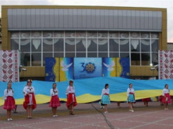 Дівчата й хлопці в українському традиційному вбранні розгортають великий синьо-жовтий прапор на площі перед будинком культури на тлі банера «30 років Незалежності». 