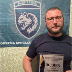 Чоловік у чорній футболці та окулярах тримає в руках рамку з грамотою «ПОДЯКА» від Новотроїцької селищної військової адміністрації Генічеського району Херсонської області, виданою Сивогривому Володимиру. На задньому плані висить темно-зелений прапор із написом «БОРИСФЕН», гаслом «ВІДВАГА І МІЦЬ» та гербом із зображенням рисі. 