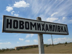 Прямокутний дорожній вказівник на залізобетонній опорі з назвою села «НОВОМИХАЙЛІВКА», написаною великими чорними літерами на білому тлі. Знаки встановлено біля дороги на тлі сільського пейзажу, полів та блакитного неба з білими хмарами. 